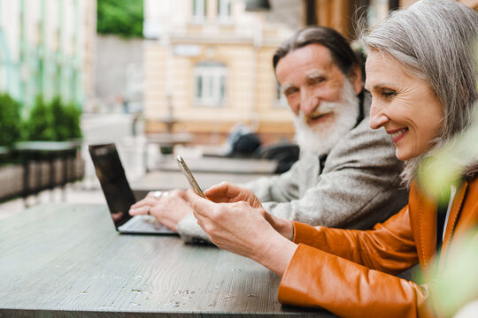 White senior couple smiling and using gadgets while sitting in cafe