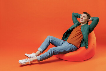 Full size body length fun young brunet man 20s wears red t-shirt green jacket sit in bag chair sleep rest keep eyes closed isolated on plain orange background studio portrait. People emotions concept.