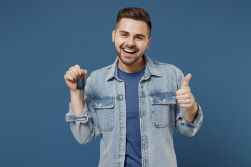 Cheery laughing satisfied handsome young brunet man 20s wears denim jacket hold key ring show thumb up like gesture isolated on dark blue background studio portrait. People emotions lifestyle concept.