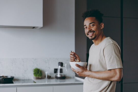Young happy smiling bachelor african american man in casual clothes eat breakfast muesli cereals with milk fruit in bowl prepare cook food in light kitchen at home alone indoor. Healthy diet concept