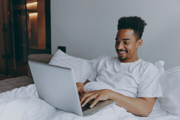 Young smiling happy fun african american man 20s wear nightwear hold use work on laptop pc computer typing message lying in bed rest relax spend time in bedroom lounge home in own room house wake up.