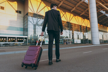 Full length back view young traveler businessman man 20s wear black classic suit walking going outside at international airport terminal with suitcase valise Air flight business trip lifestyle concept