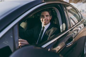 Side profile view young driver confident smiling businessman man 20s wear black dinner suit driving car taxi hold steering wheel look camera Vehicle transport traffic lifestyle business trip concept