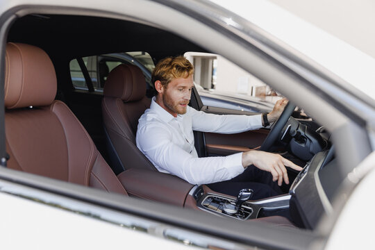 Man customer buyer male client wearing white shirt sitting in car salon turn on music drive chooses auto wants buy new automobile in showroom vehicle dealership store motor show indoor Sales concept