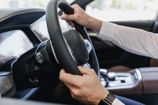 Close Up Cropped Up Photo Shot Hands Arms European Businessman Man Wearing White Shirt Sitting In BMW Brand Car Salon Driving Hold Steering Wheel Automobile Modern Vehicle. Car Sales Driver Concept