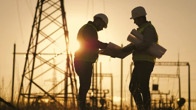 Teamwork industry a concept. Two engineers electric experts in protective helmets discussing the construction of a power line. Silhouette Of Engineers Standing On Field With Electricity Towers.