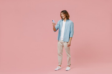 Full length young happy satisfied cool man with long curly hair wear blue shirt white t-shirt hold in hand use mobile cell phone isolated on pastel plain pink color wall background studio portrait