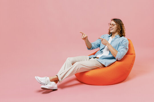 Full length young happy man with long curly hair in blue shirt white t-shirt glasses sit in bag chair point index finger aside on workspace area isolated on pastel plain pink color background studio