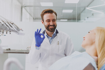 Obraz premium Young smiling caucasian man dentist doctor in white gown show okay gesture consulting patient woman sitting at dentist office chair indoor cabinet near stomatologist Healthcare enamel aesthetreatment