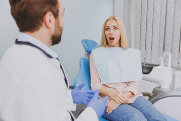 Young man dentist doctor in white gown talk speaking with patient consulting shocked woman sit at dentist office chair indoor cabinet near stomatologist wait medical cure Healthcare enamel treatment