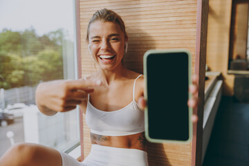 Close up young happy sportswoman woman in white sportswear earphones listen music point finger on mobile cell phone with blank screen area sit on windowsill of gym rest inside People lifestyle concept