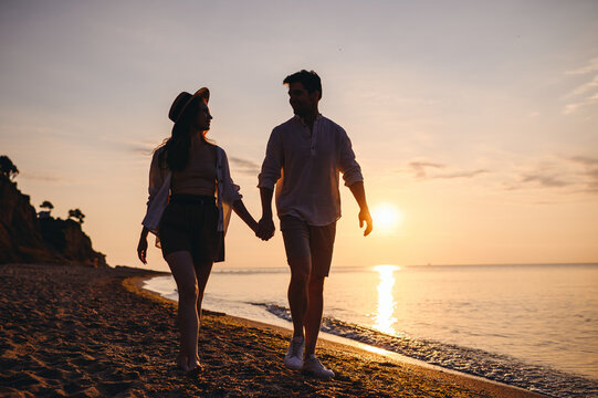 Full body lower silhouette young couple two friends family man woman in casual clothes hold hand look to each other walk together at sunrise over sea beach ocean outdoor seaside in summer day evening.