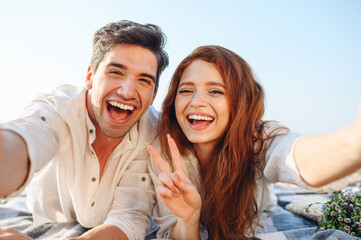 Fototapeta premium Close up young couple two family man woman in white clothes lying on plaid do selfie shot pov on mobile phone show v-sign rest together at sunrise over sea beach ocean outdoor seaside in summer day