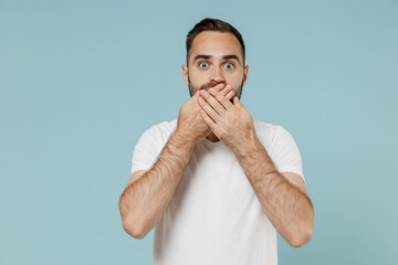 Young shocked astonished surprised man 20s wearing basic casual white t-shirt covering mouth with hands isolated on plain pastel light blue color background studio portrait. People lifestyle concept.