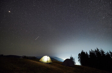 Night camping in the mountains. Two tents, one of which illuminated, set up on mountain meadow. Panoramic view of evening sky with multiple stars.