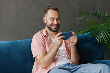 Young gambling happy man in casual clothes using play racing app on mobile cell phone gadget smartphone for pc video games sitting on blue sofa at home flat indoors rest relax on weekends free time - obrazy, fototapety, plakaty