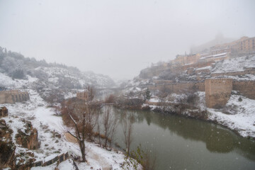 City landscape of snowy old city and castle with clouds and fog; View of the Tajo river, Alc&aacute;zar and Alc&aacute;ntara bridge from Toledo during the storm Filomena, World Heritage Site, Spain. Horizontal view