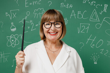 Smiling clever teacher mature elderly lady woman 55 wear shirt glasses holding pointer in hand isolated on green wall chalk blackboard background studio. Education in high school September 1 concept