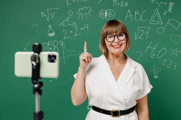 Teacher mature elderly woman 55 wear shirt glasses work with mobile cell phone on tripod quarantine hold index finger up with great new idea isolated on green wall chalk blackboard background studio.