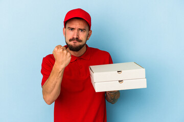 Young caucasian delivery man with tattoos holding pizzas isolated on blue background  showing fist to camera, aggressive facial expression.