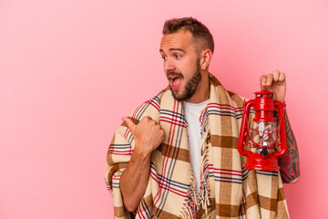 Young caucasian man with tattoos holding vintage lantern isolated on pink background  points with thumb finger away, laughing and carefree.
