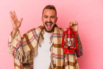 Young caucasian man with tattoos holding vintage lantern isolated on pink background  receiving a pleasant surprise, excited and raising hands.