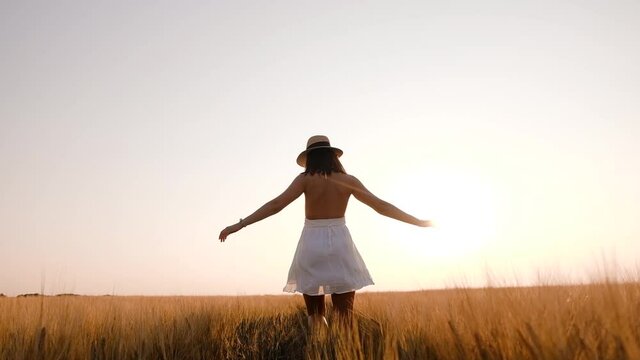 Happy free young woman dancing and spinning around in slow motion across field, touching ears of wheat with her hand. Wheat field on sunset background