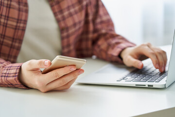 Unrecognizable woman surfing mobile phone while entering data on computer. Cybersecurity awareness and data managment in digitally-connected world. 2fa verification and authentication