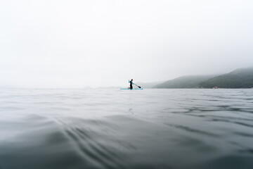 Naklejka premium Fitness woman standing on surfboard enjoying supsurfing with paddle at endless sea water fog