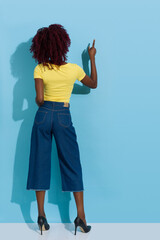 Young Black Woman Is Standing And Pointing At Blue Sunny Wall. Rear View.