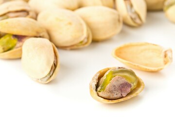 Close up of single, open pistachio in front of heap of salted, roasted green pistachio nuts snack over white background, healthy food snack