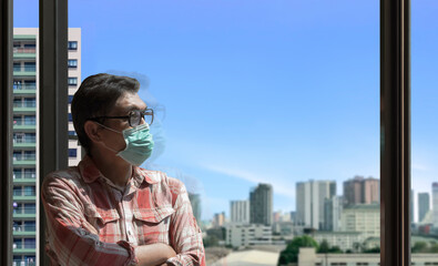 Fototapeta na wymiar Asian man wearing protective mask and looking out of glass window at home with blurred city view background, He hoped that the Coronavirus outbreak problem will end soon