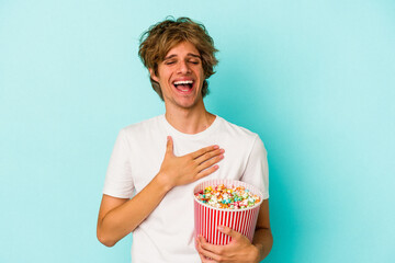 Young caucasian man with makeup holding popcorn isolated on blue background  laughs out loudly keeping hand on chest.