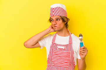 Young ice cream maker man with makeup holding spoon isolated on yellow background  touching back of head, thinking and making a choice.