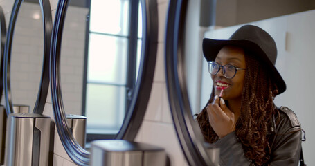 Afro-american young woman apply lipstick looking into mirror in public toilet