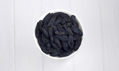 Dark blue raisins in a white bowl on a light wooden surface. Organic sugar free dried grapes.