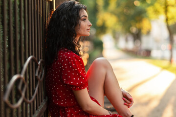 Beautiful young woman with curly hair in a red vintage dress sits near the fence in the park at...