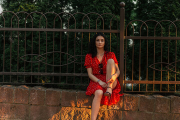 Fashionable beautiful woman model in red dress sits near a fence in the city