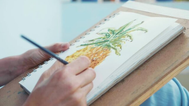 Close-up of female hand with paintbrush is drawing pineapple in sketchbook by watercolors
