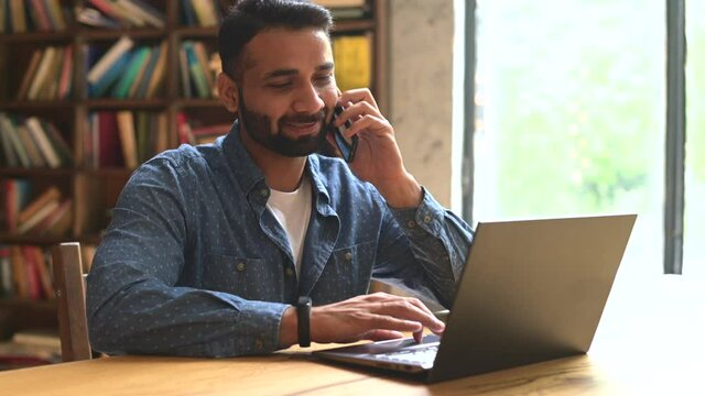Mid adult indian man working in his home office, using smartphone and laptop for communication with customers or colleagues, smiling multiracial eastern guy talking phone watching on computer screen