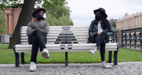 African young man and woman in safety mask sitting on bench in park during coronavirus outbreak