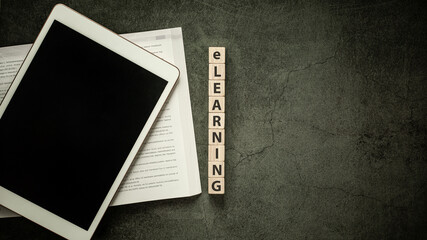 Education e-learning concept, Blank screen tablet with notebook and word e-learning on wood cube block on black table background, Flat lay top view copy space.