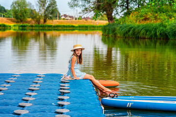 A cute little girl in dress is sitting on the edge of the boat dock by the lake. Beautiful summer landscape