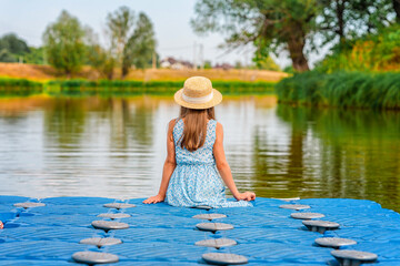 A cute little girl in dress is sitting on the edge of the boat dock by the lake. Beautiful summer...