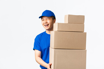Online shopping, fast shipping concept. Cheeky smiling asian courier in blue uniform holding boxes with client orders, wink friendly at customer, provide fast delivery, standing white background - obrazy, fototapety, plakaty