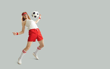 Fototapeta na wymiar Excited nerdy male sports fan having fun with a football. Happy funny young man in white and red activewear kicking away soccer ball on light gray background with empty space for advertising text