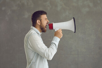 Casual adult man shouting in megaphone loudspeaker studio shot with copy space. Displeased nervous businessman freelancer loudly screaming at bullhorn. Announcement and advertisement concept