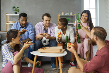 Casual group of happy young diverse friends relaxing on floor and sofa, eating pizza slices from...