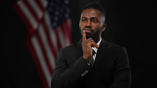 Troubled thoughtful African American elegant man in suit waiting for public speaking at black background with USA flag. Portrait of anxious male candidate getting ready for debates thinking