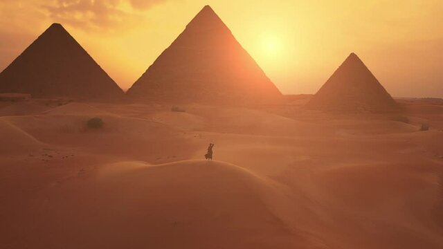 Female traveller looks to the silhouette of pyramids at the sunset in the desert. Aerial shot.